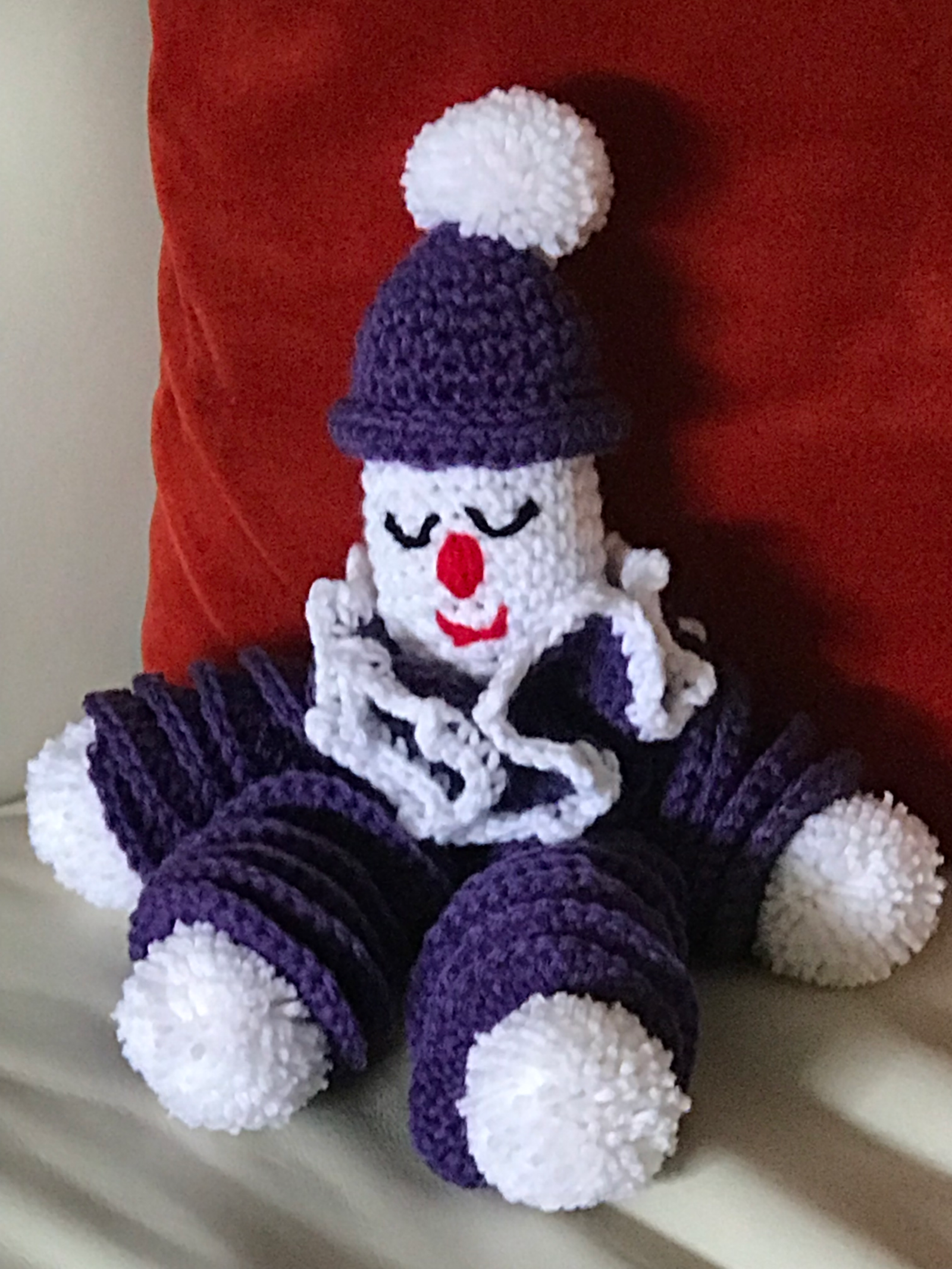 Clownie the Crocheted Clown Doll — a purple and white crocheted clown with pom-poms, seated against a red cushion